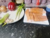 Fresh ingredients for a salmon vegetable medley, including salmon fillets, spring onions, garlic, red bell pepper, and onion arranged on a dark kitchen counter.
