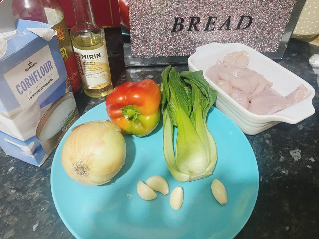 Fresh stir-fry ingredients including chicken, onion, garlic, bell pepper, and pak choi displayed on a blue plate.