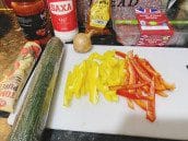 Colorful fresh ingredients including diced yellow and red bell peppers, a cucumber, an onion, minced beef, and essential seasonings displayed on a kitchen counter.