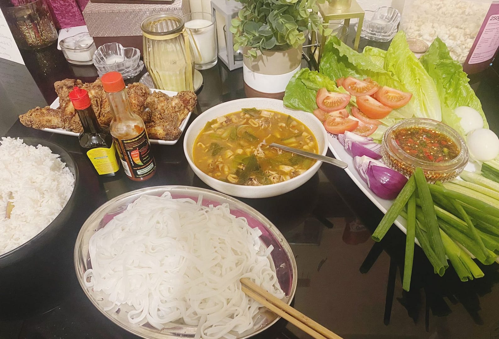 A vibrant Asian fusion meal with crispy chicken wings, rice noodles, seafood broth, and a fresh salad platter arranged on a dinner table.