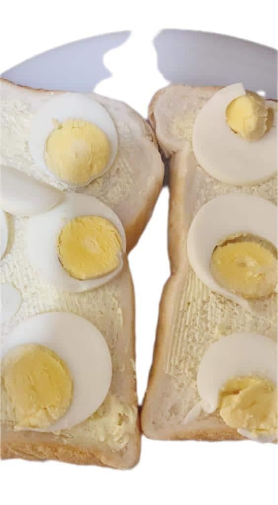 Sliced boiled eggs placed beside two buttered bread slices on a dark prep surface, ready for sandwich assembly.