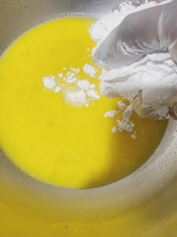 Flour is being added by hand into a bowl of yellow dough mixture.