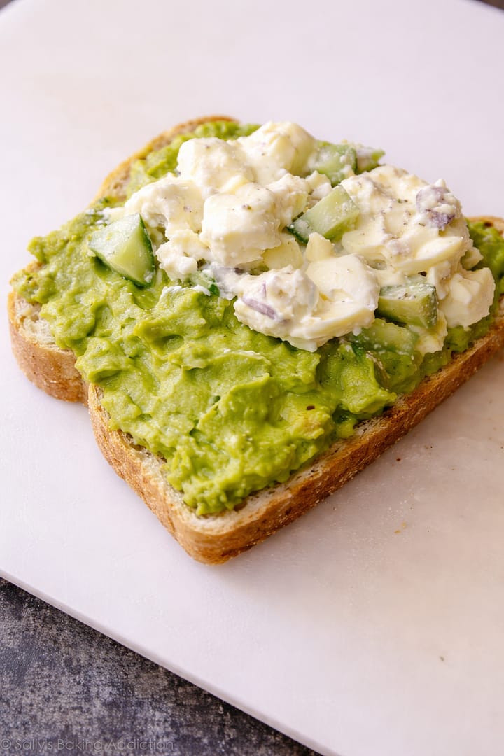 Avocado toast topped with creamy egg salad, cucumber, and onion on toasted bread