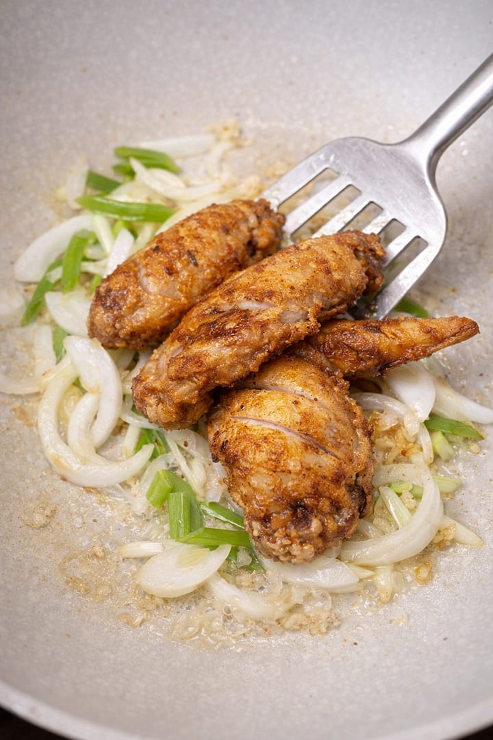 Golden fried chicken wings being added to sautéed onions and spring onions in a pan