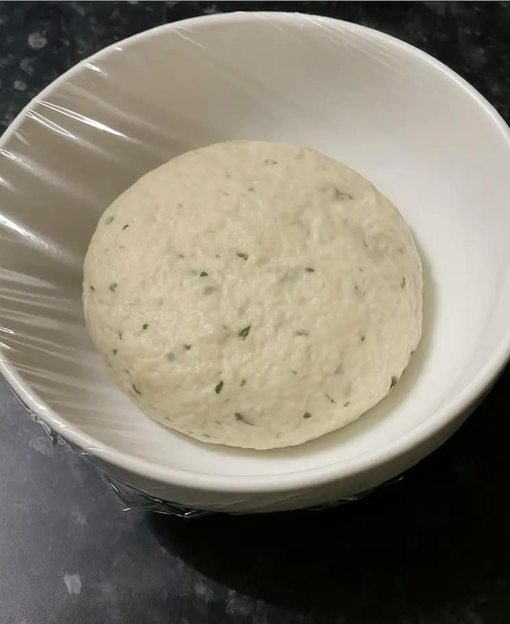 Smooth naan dough resting in a clean bowl covered with cling film, ready to rise.