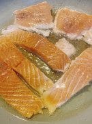 Raw salmon fillets sizzling in hot oil inside a frying pan, beginning to sear.