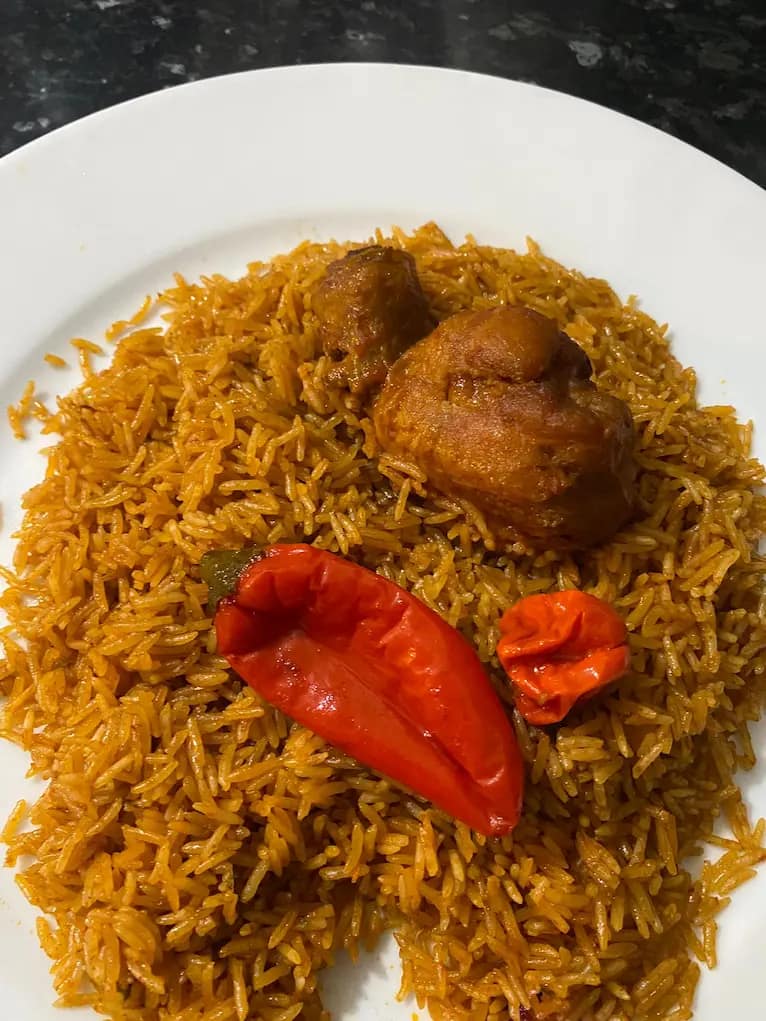 A serving plate with bright red Jollof rice topped with golden chicken drumsticks, garnished with herbs.