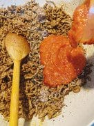 Tomato sauce being poured into browned minced beef inside a pot, with a wooden spoon beside it.