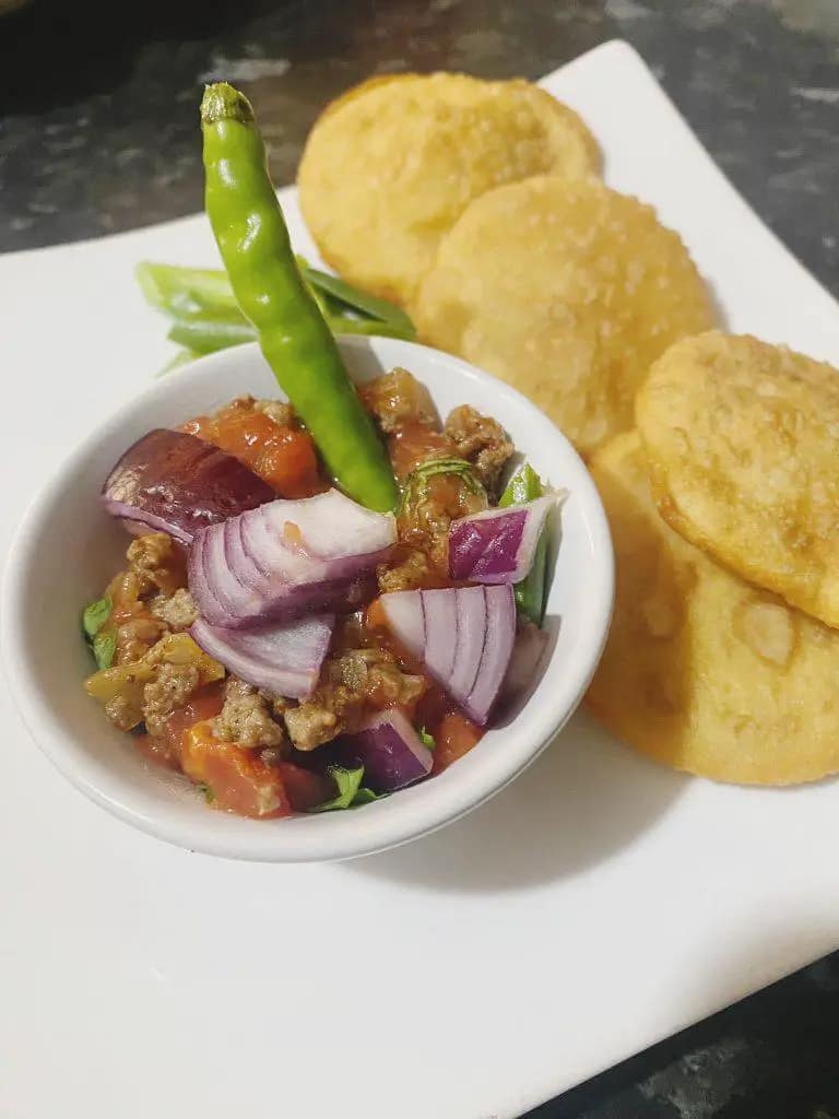 A bowl of savory minced beef garnished with red onions and green chili, served alongside golden fried dough rounds on a white plate.