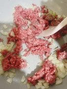 Minced beef cooking with sautéed onions in a pot, stirred with a wooden spatula for a savory base.