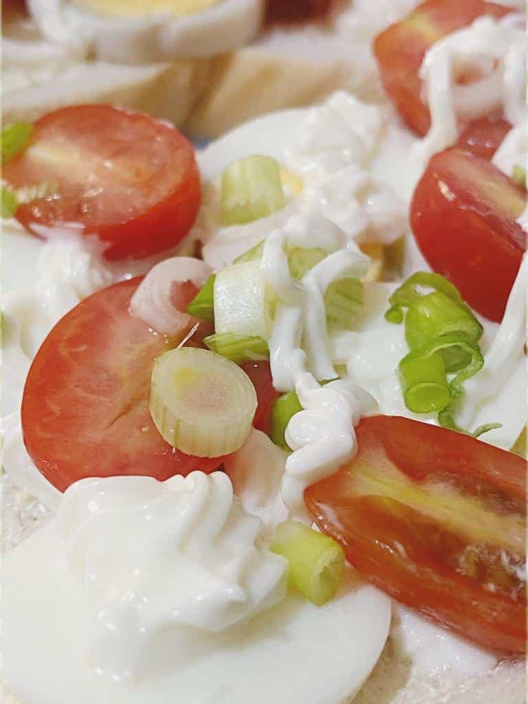 A close-up view of a golden egg and mayo breakfast sandwich, sliced in half, showing layers of boiled egg, tomato, and chopped spring onions on buttered bread.