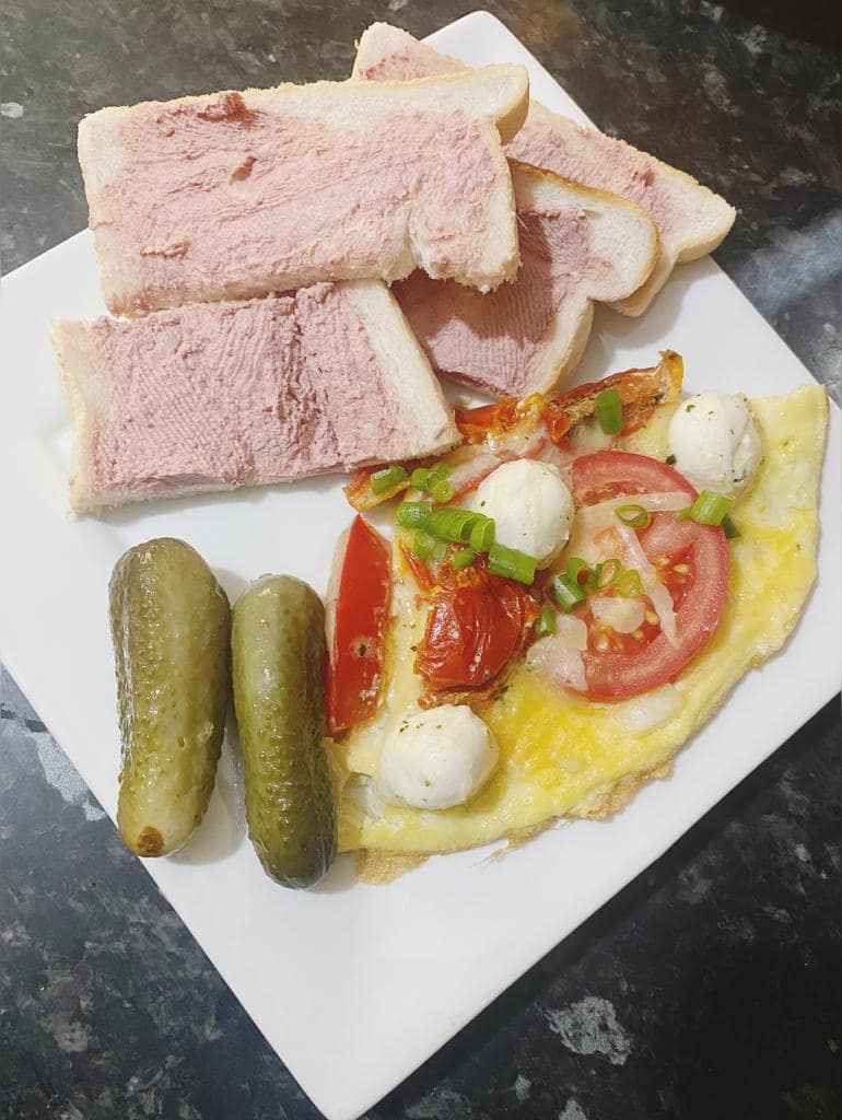 Cheesy tomato omelette served with pΓ’tΓ© on white bread and two pickles on a white plate.