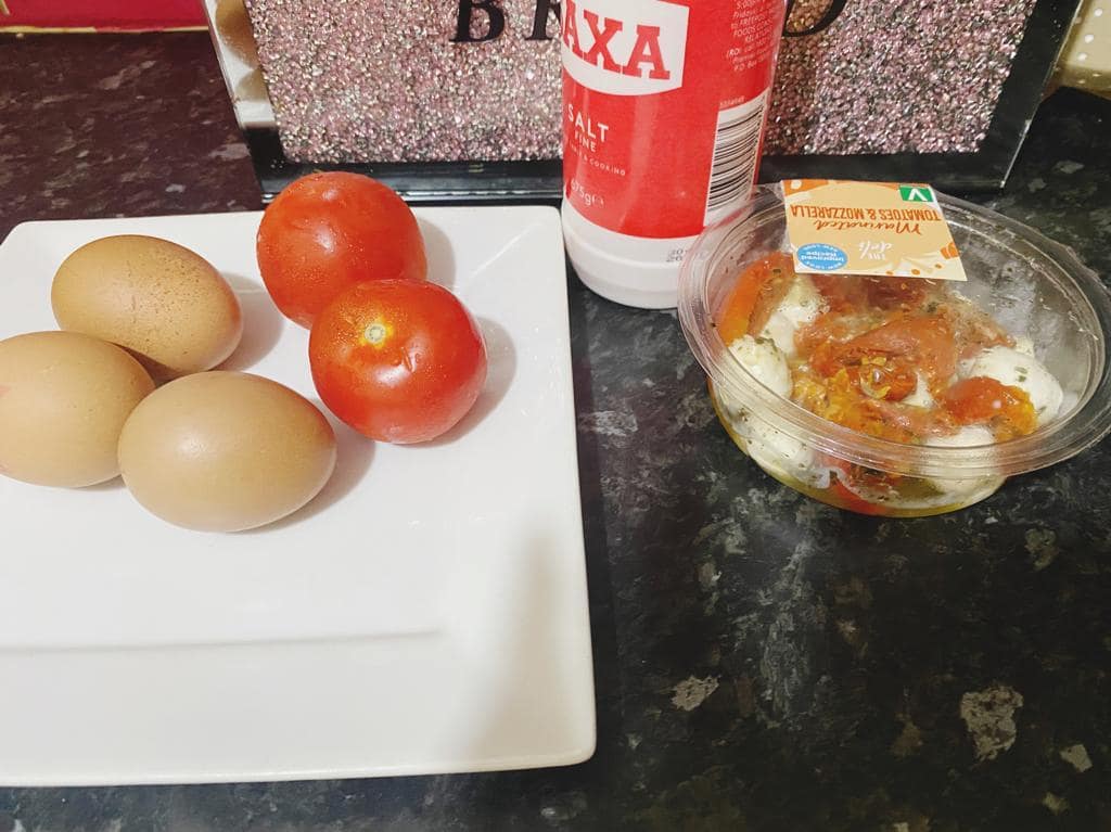 Ingredients on a kitchen countertop: a plate with four brown eggs and two tomatoes, a container of seasoned mozzarella and tomatoes, and a salt container.